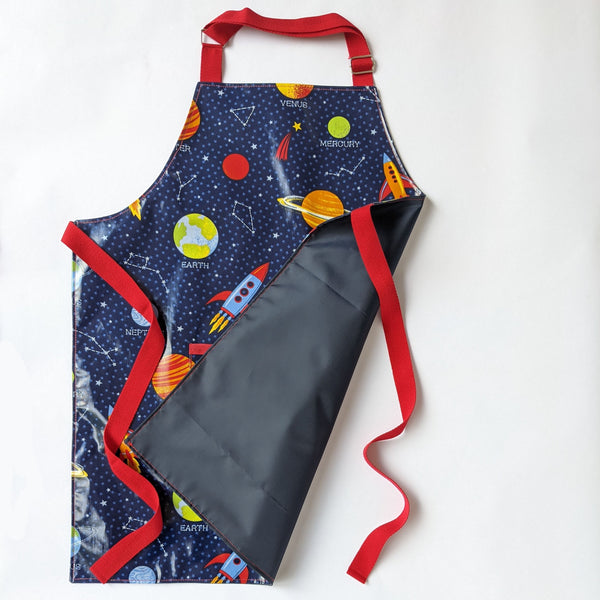 Large Kids, Wipeable, Space Oilcloth Apron with front pocket,  Red Adjustable neck band and red ties and trim. Water resistant navy lining. 